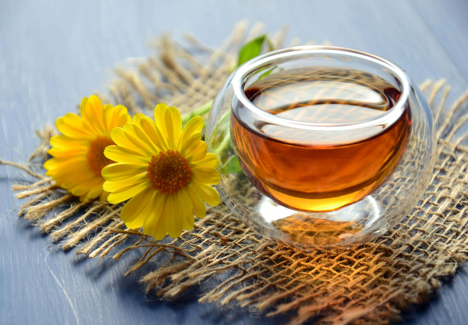 A clear glass tea cup filled with a golden tea on a bamboo mat with fresh flowers next to it.